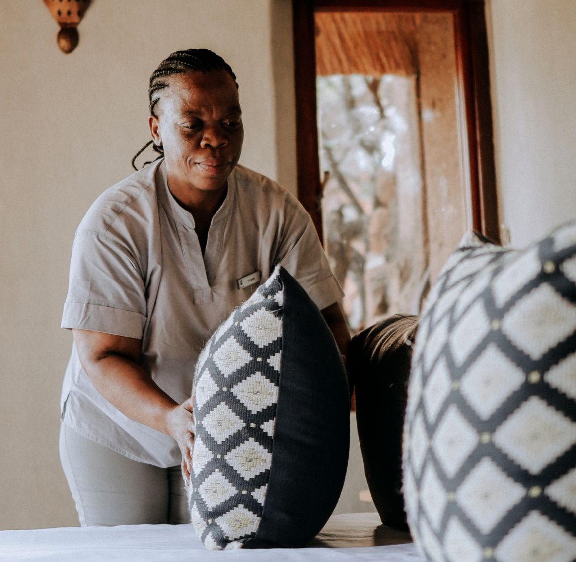 Light Housekeeping Caregiver tidying up decorative pillows on a bed in a warmly lit room.