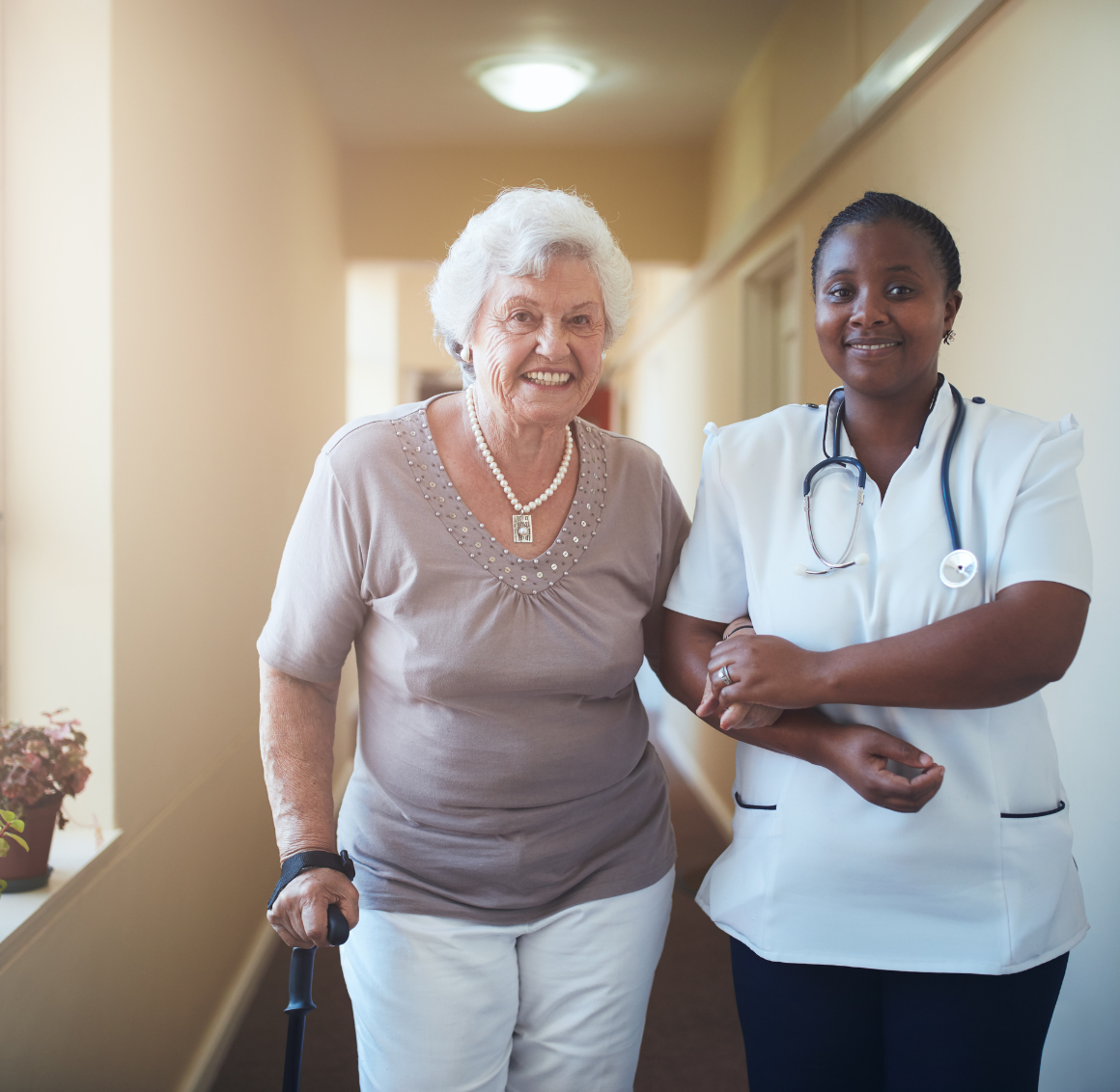 Live-in Care Smiling senior woman walking arm-in-arm with a nurse in a hallway.