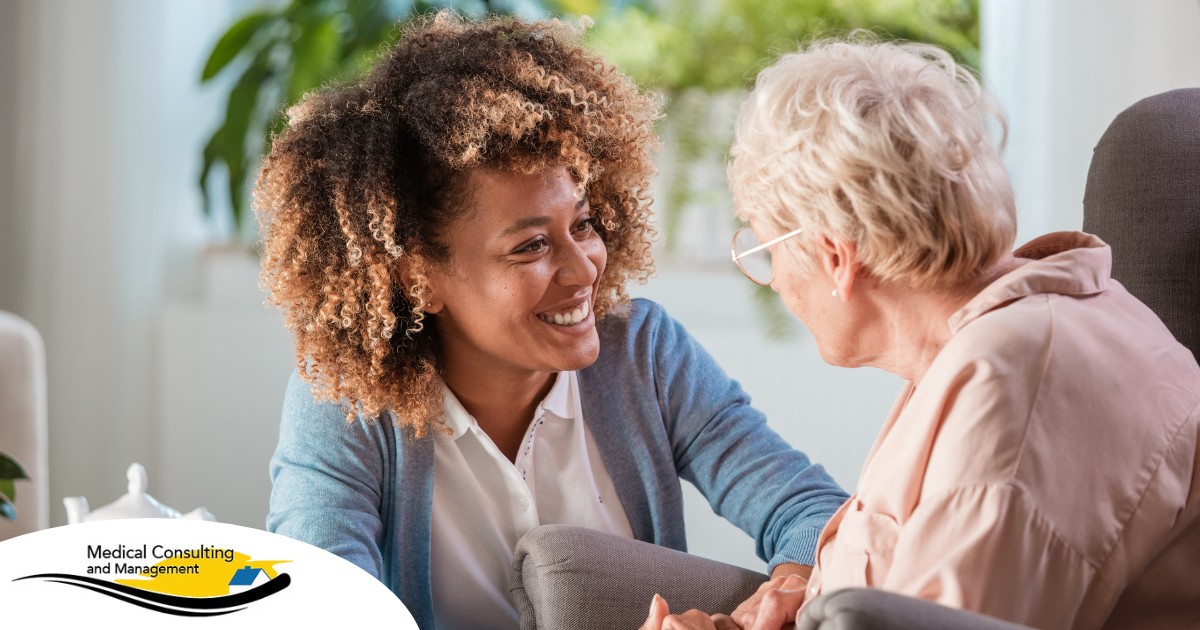 A caregiver smiles at an older client in a chair, representing the joy that can come from a healthy work-life balance.