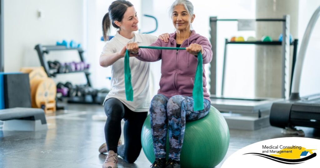 A care provider helps an older woman exercise with a resistance band and an exercise ball, representing how exercise can help with senior fall prevention.