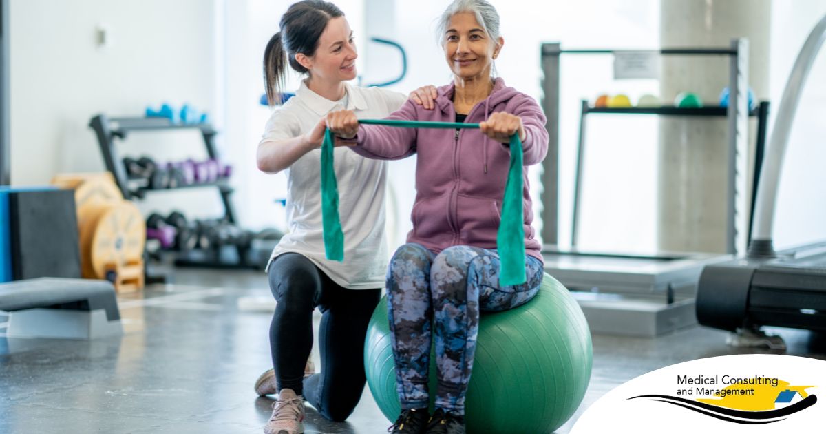 A care provider helps an older woman exercise with a resistance band and an exercise ball, representing how exercise can help with senior fall prevention.