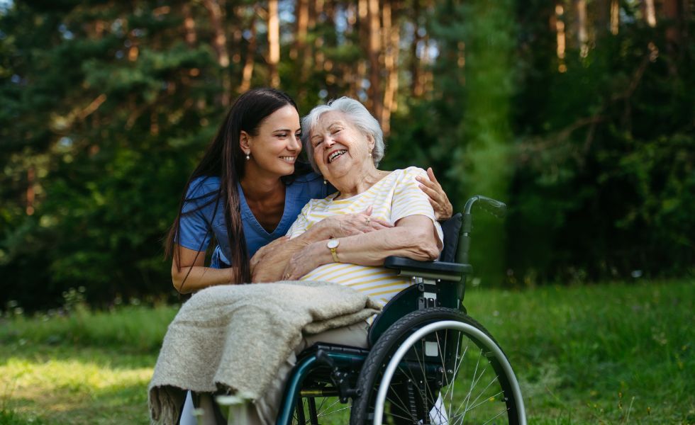 Caregiver smiling and embracing an elderly woman in a wheelchair outdoors, representing Alzheimer’s & Dementia Care in Rockville.